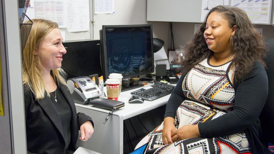 two women sit and talk in office cubicle