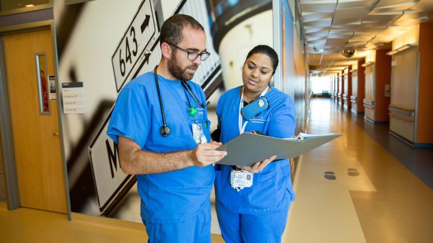 Two nurses look at a clipboard in the hallway